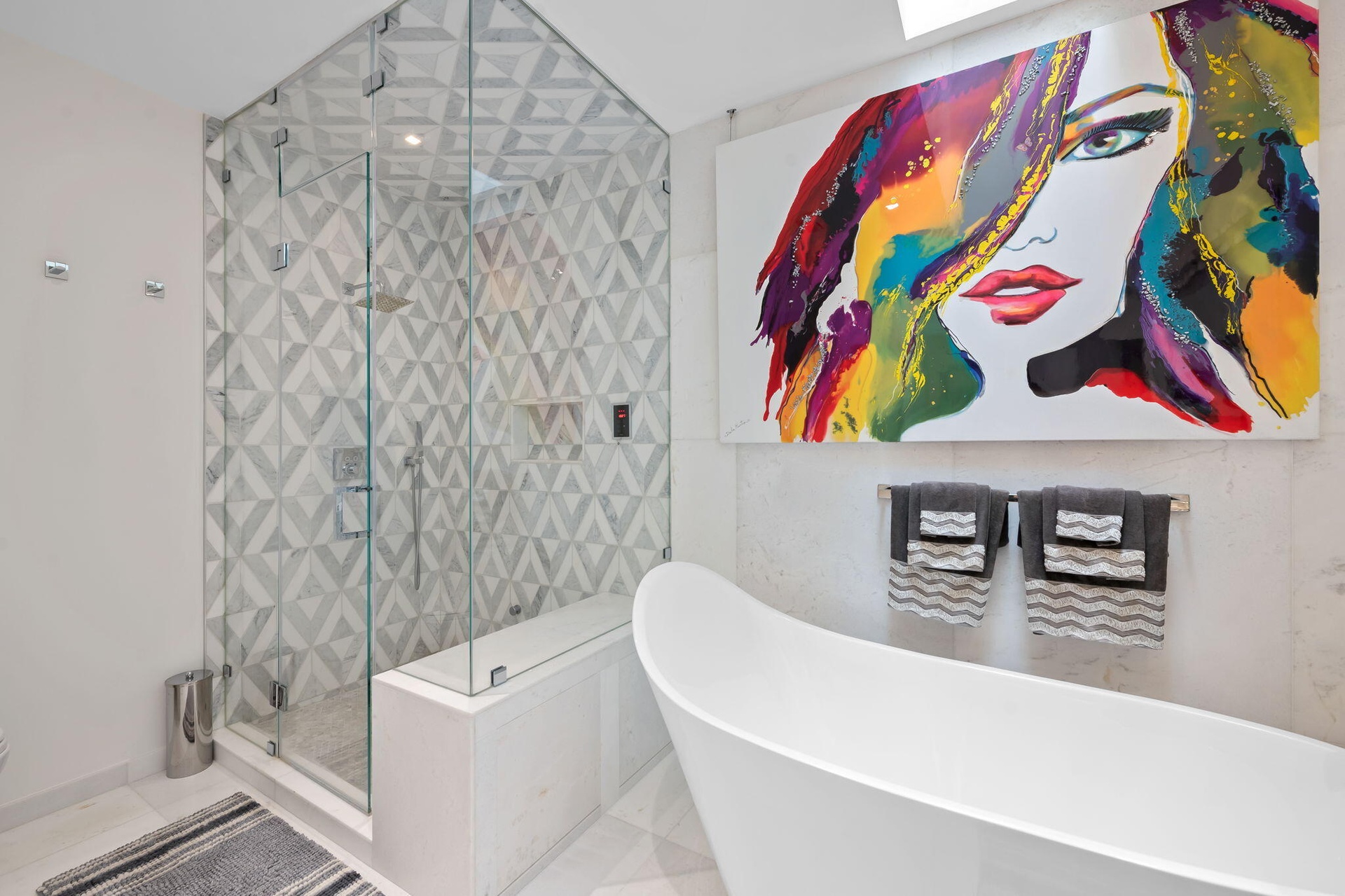Freestanding soaking tub, glass-enclosed walk-in steam shower with geometric patterned tile, and polished natural stone wall surfacing. Newly remodele