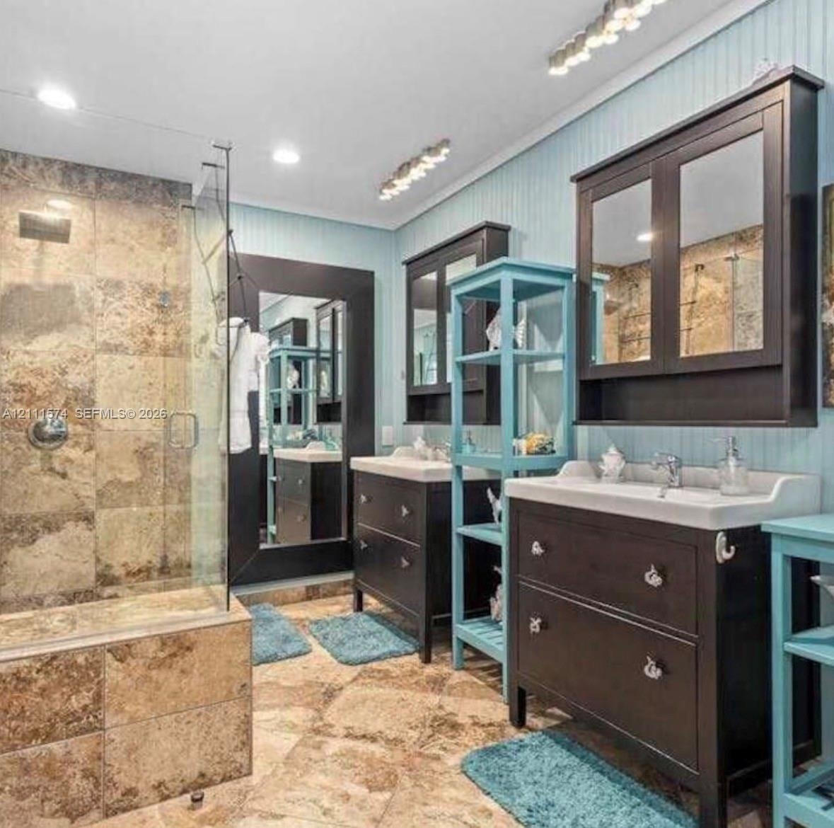 Bathroom featuring a walk-in shower with stone tile surround, dual dark-finish vanities with integrated cabinetry, beadboard wall paneling, and stone 