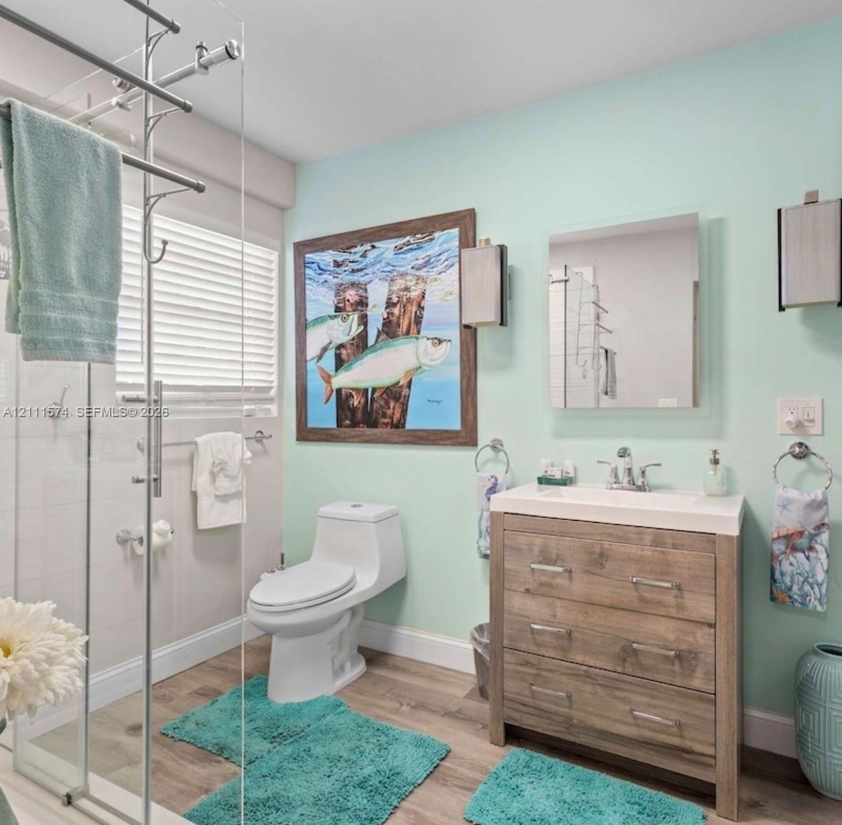 Bathroom featuring wood-finish flooring, a wood-grain vanity with a white countertop, a frameless glass-enclosed shower, and wall-mounted rectangular 