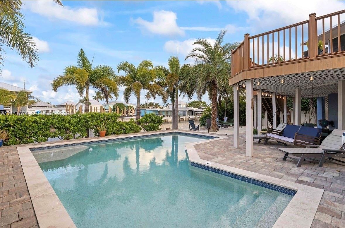In-ground swimming pool with a stone paver deck, elevated wood-tone balcony, and covered lower-level patio area