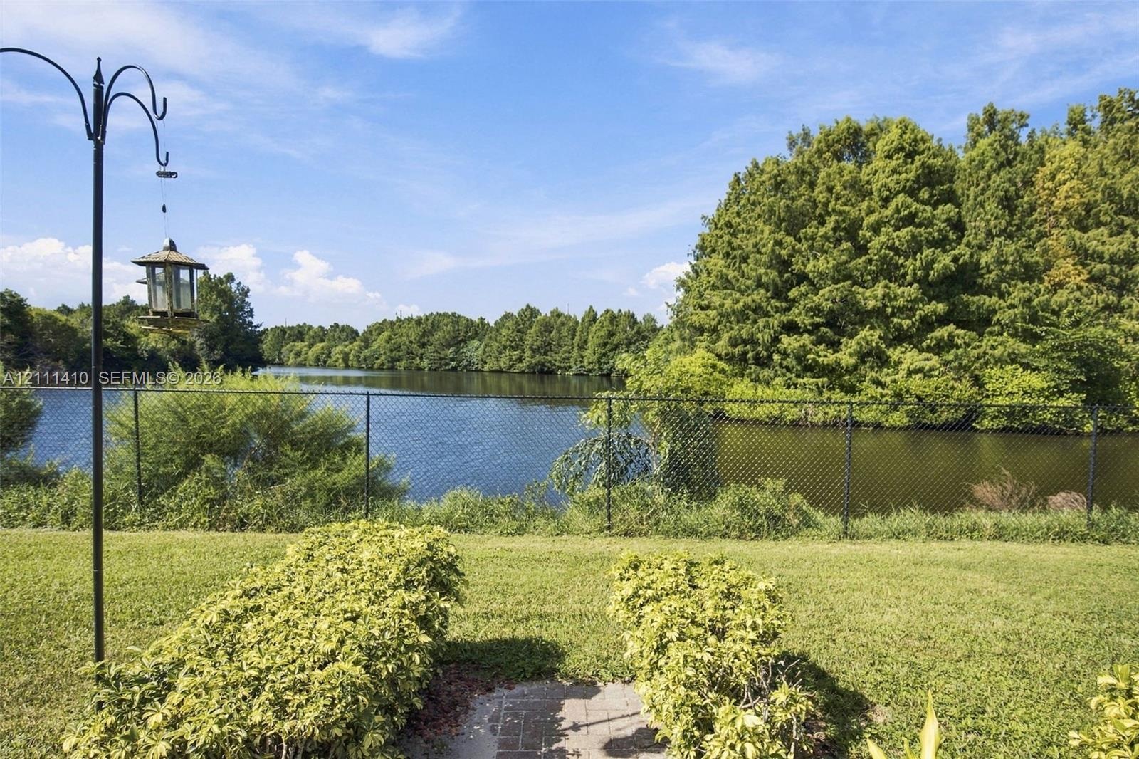 Backyard featuring a manicured lawn, established shrubbery, a black metal chain-link fence, and direct water frontage with dense natural perimeter tre