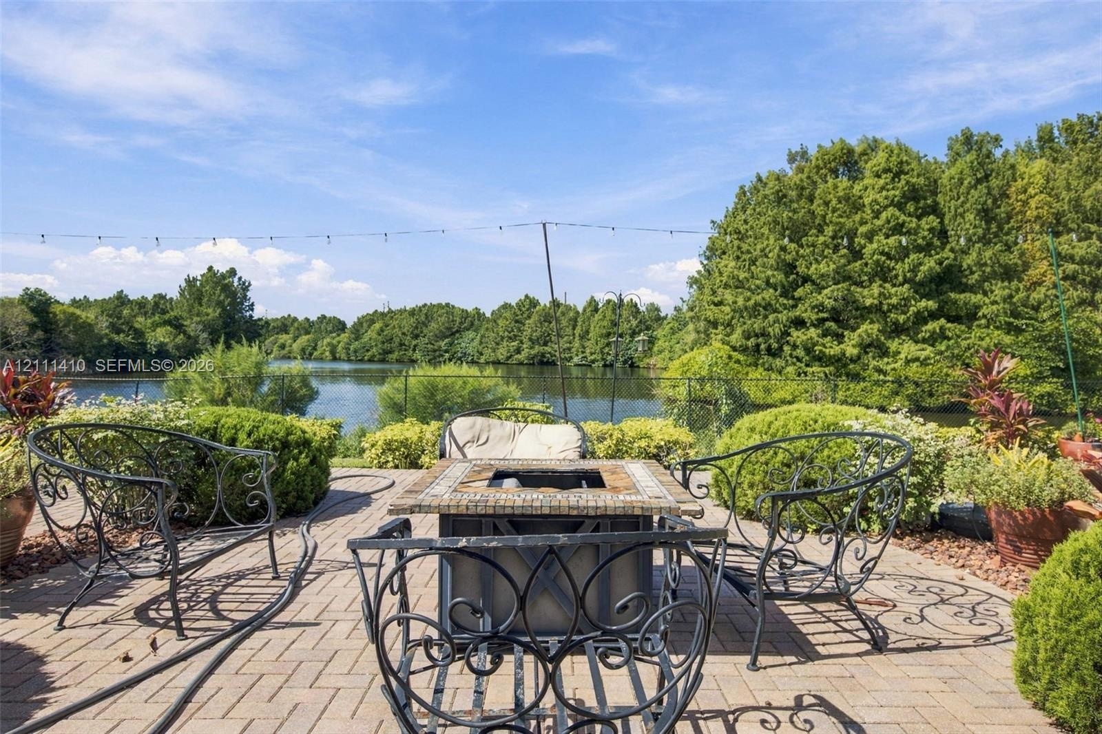 Paver patio featuring a decorative stone-tiled fire pit and perimeter landscaping with mature trees overlooking a waterfront backdrop