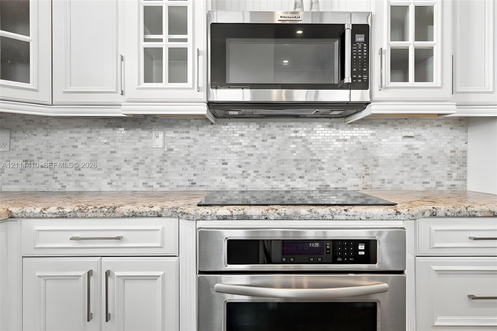 White shaker-style cabinetry with glass-front uppers, stone-finish countertops, mosaic tile backsplash, and stainless steel built-in appliances includ