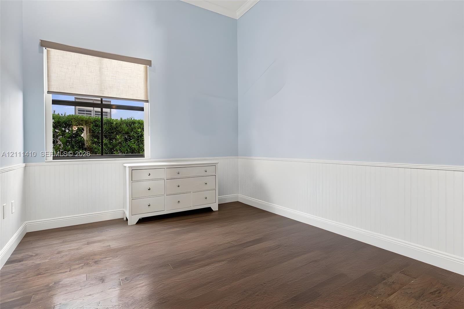 Interior room featuring wood-finish flooring, white beadboard wainscoting, and a single-pane window with a light-filtering roller shade