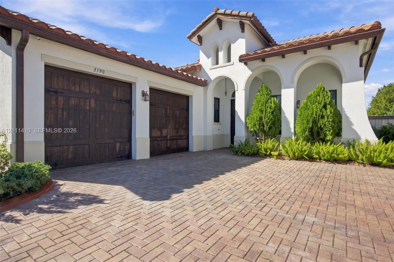 Stucco exterior featuring arched entryways, a terracotta-style tile roof, dual wood-finish garage doors, and a herringbone-patterned paver driveway