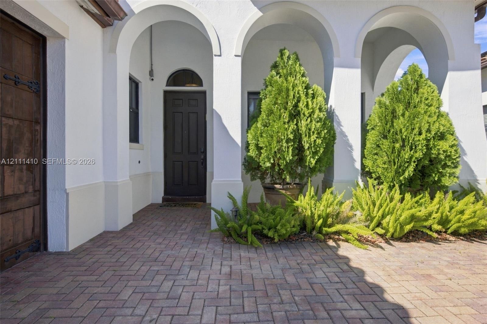 Covered entryway featuring arched architectural detailing, herringbone-patterned brick pavers, a solid wood-finish entry door with metal hardware, and
