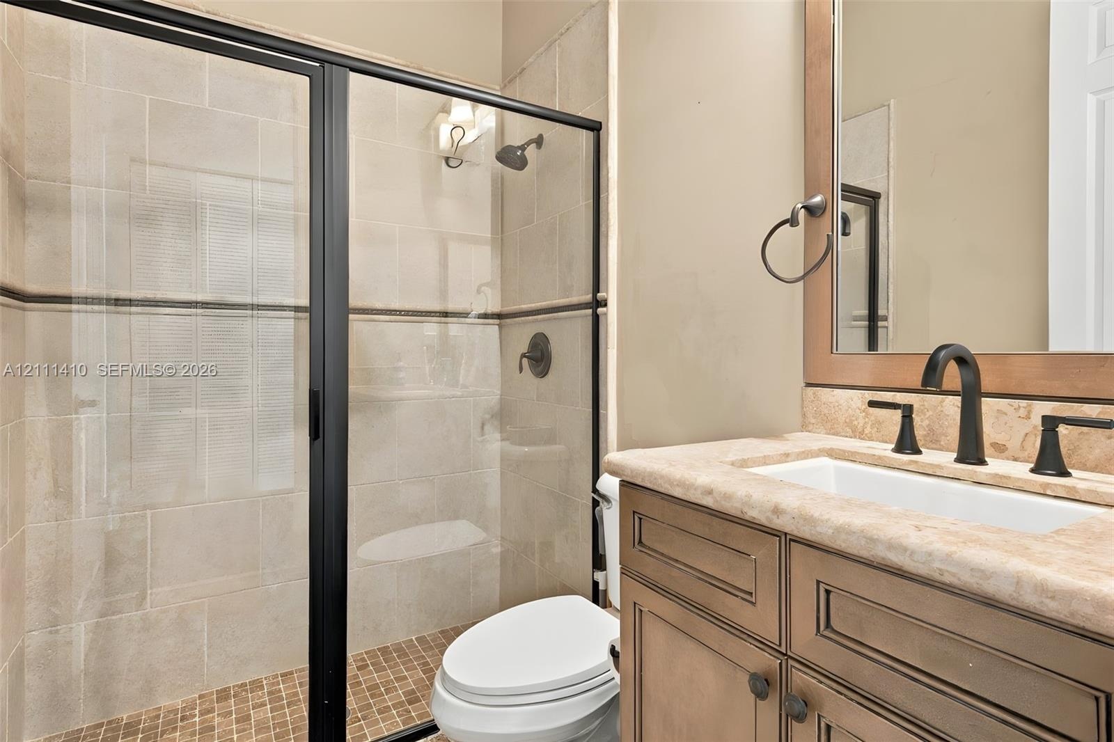 Shower with glass enclosure and black frame, vanity featuring a stone countertop and rectangular undermount sink, dark-finished hardware, and tile wal