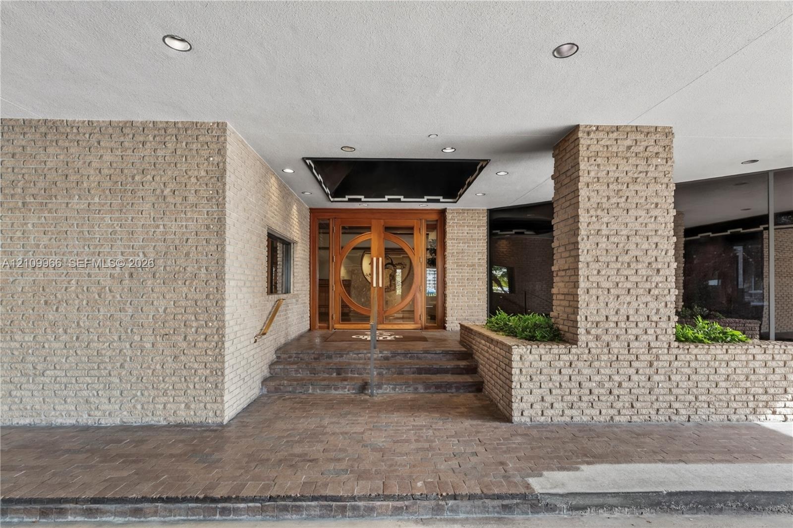 Building Exterior entrance featuring light-toned brick masonry, stone steps, dual wood-framed circular glass doors, recessed ceiling lighting, and int