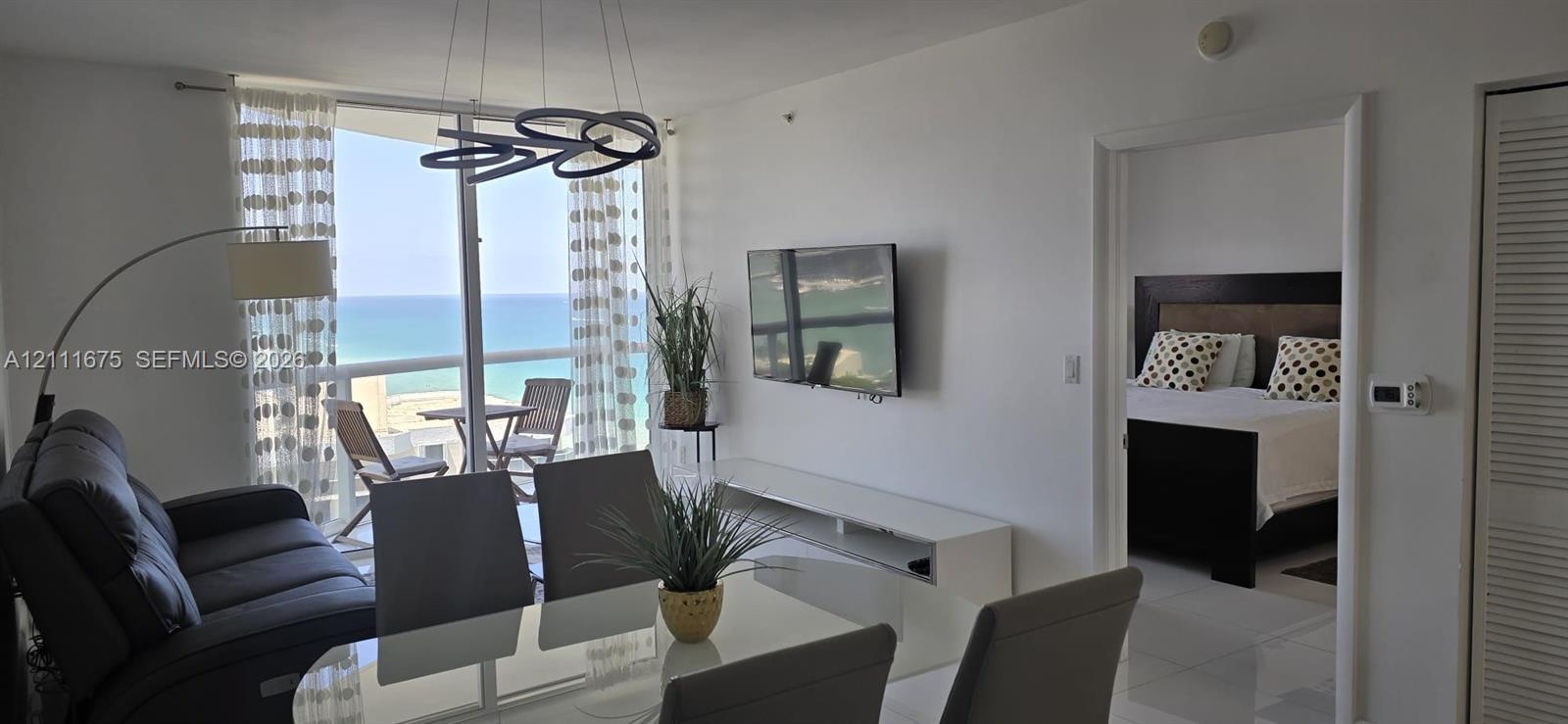 Floor-to-ceiling glass balcony doors, sculptural overhead lighting fixture, white tile flooring, and coastal scenery