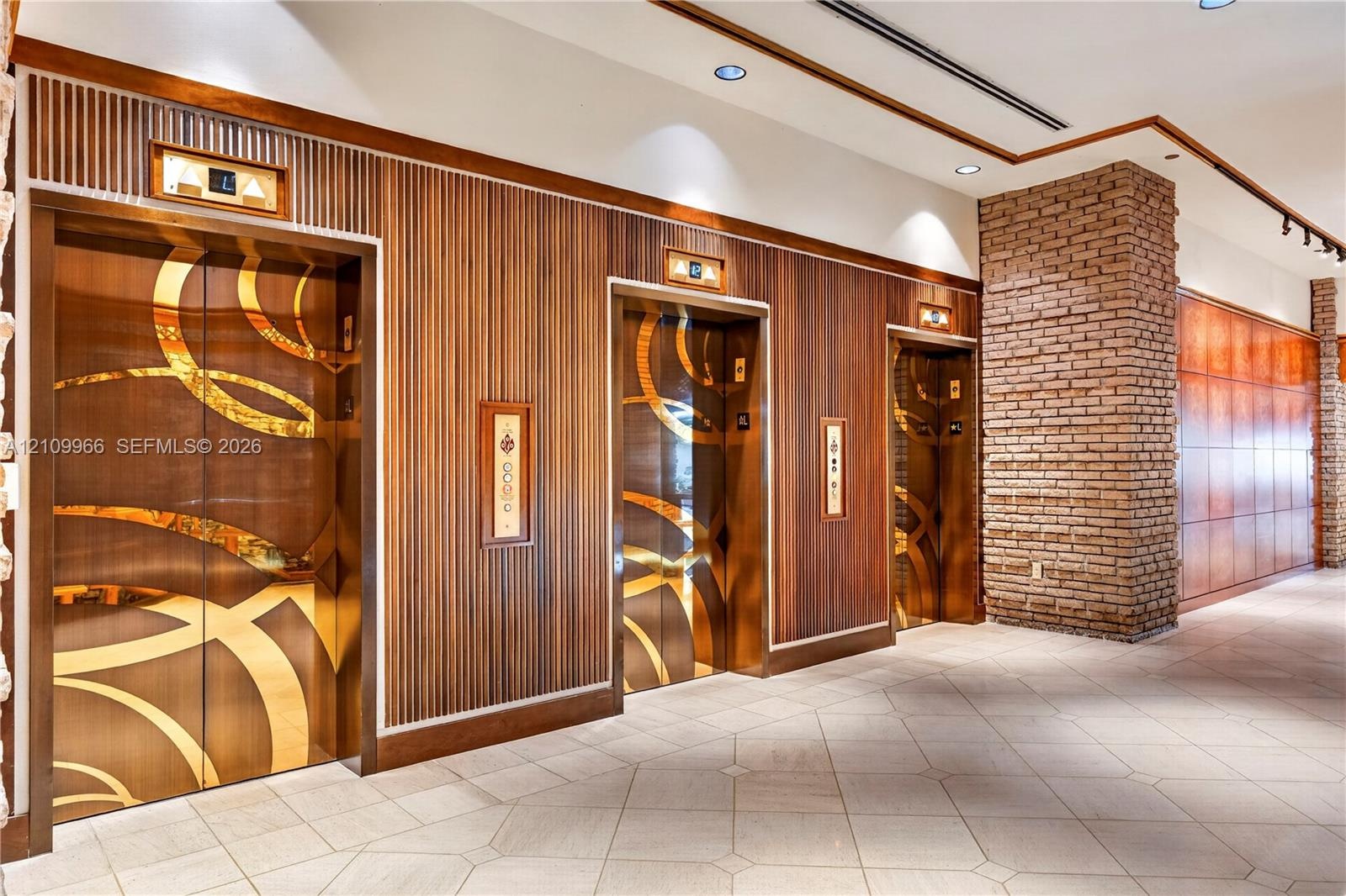Elevator lobby featuring metallic geometric door panels, slatted wood wall cladding, exposed brick masonry pillar, recessed lighting, and tile floorin