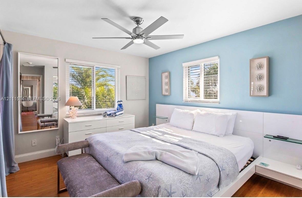 Bedroom featuring wood-finish flooring, a contemporary six-blade ceiling fan with integrated lighting, and a built-in white panel headboard with glass