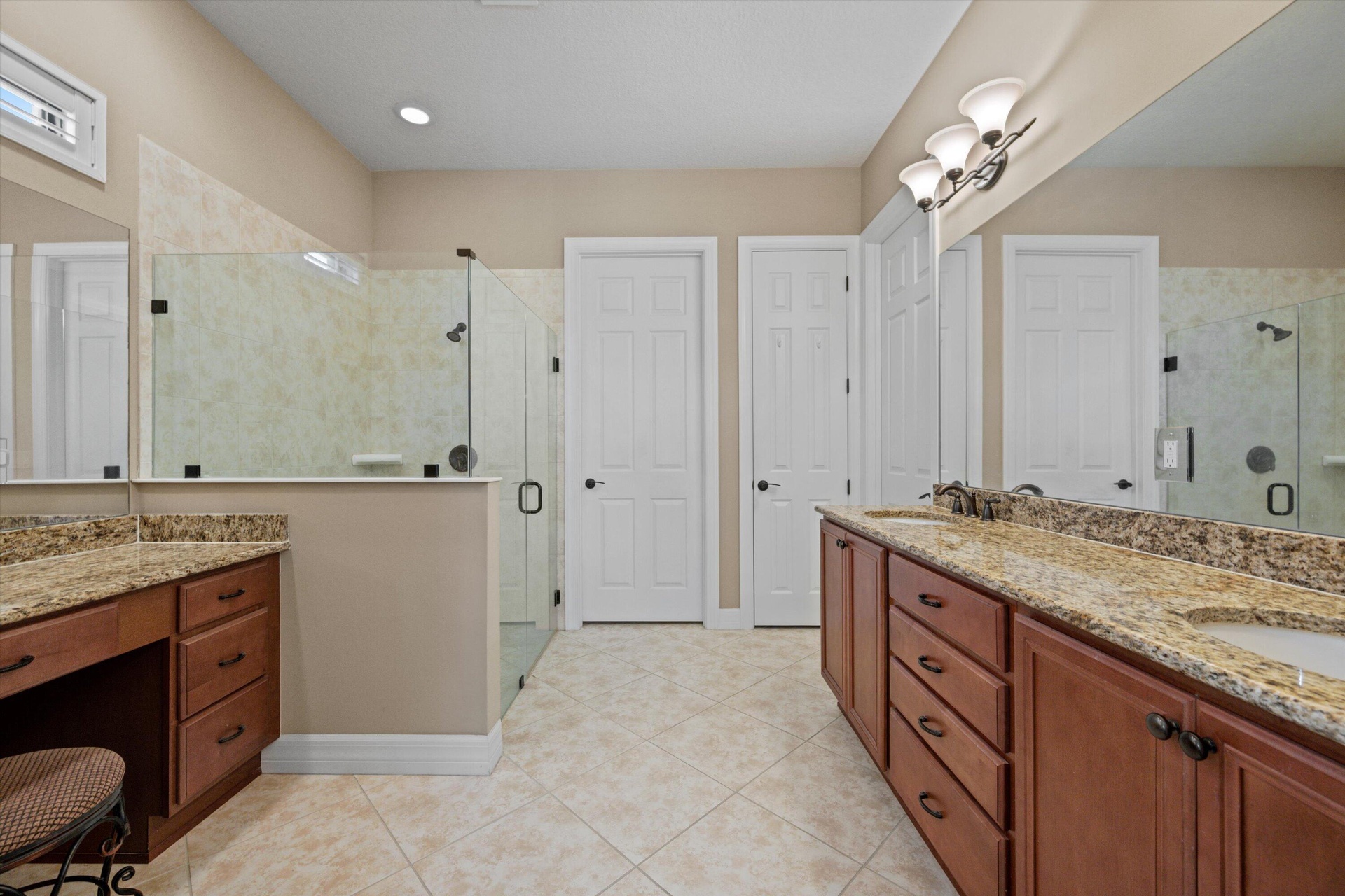 Bathroom featuring dual-sink vanity with stone-finish countertops, glass-enclosed shower, dedicated vanity seating area, and tile flooring