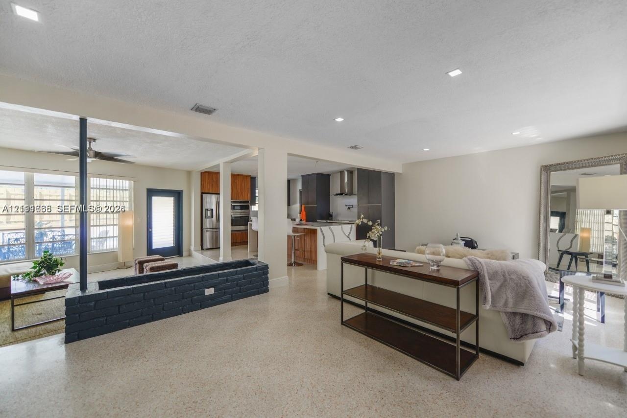 Open-concept interior featuring light-toned stone flooring, a painted brick structural partition, recessed lighting, and a kitchen equipped with dark 
