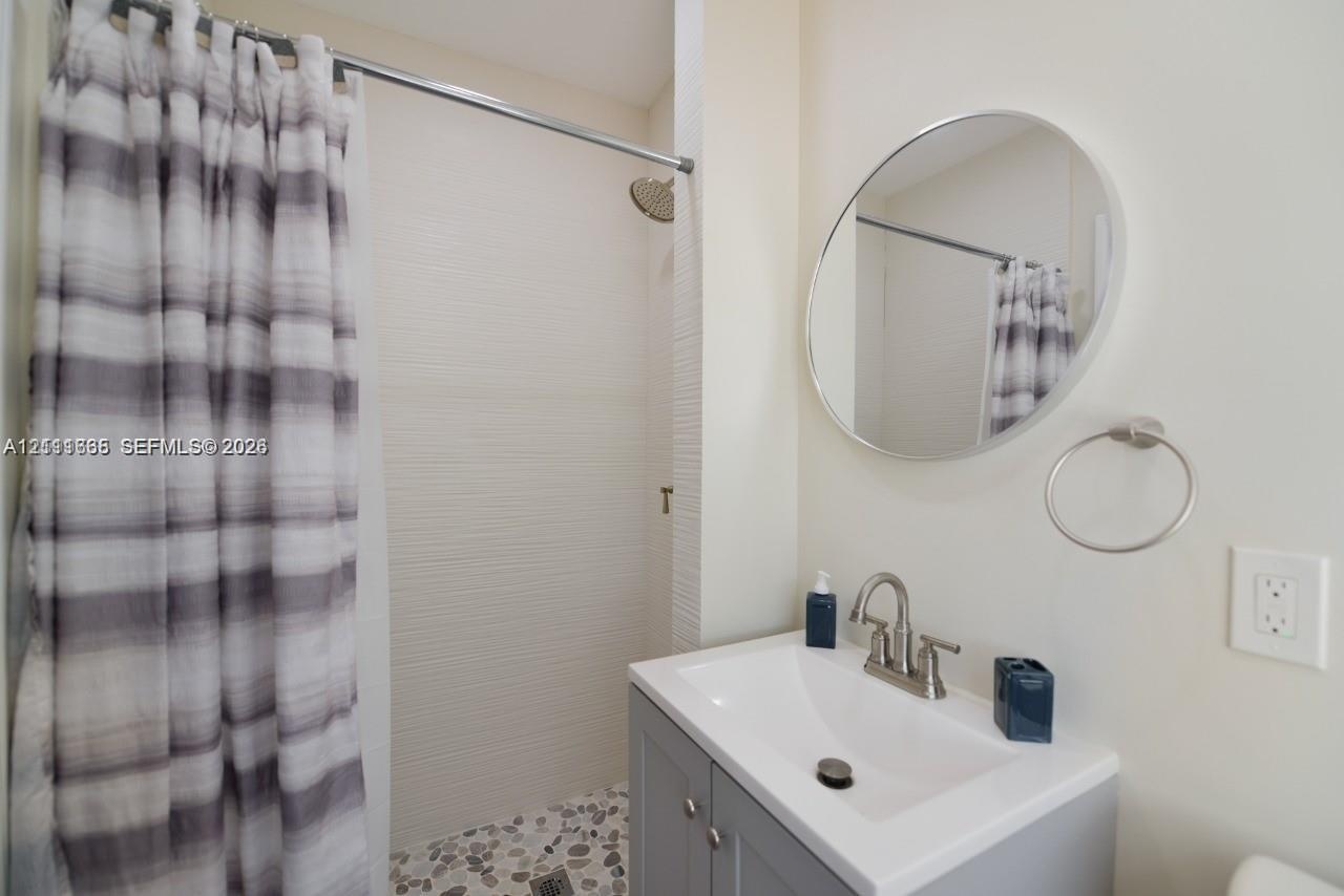 Bathroom featuring a gray vanity with an integrated basin, circular wall mirror, wall-mounted towel ring, and a tiled shower stall with a rain-style s