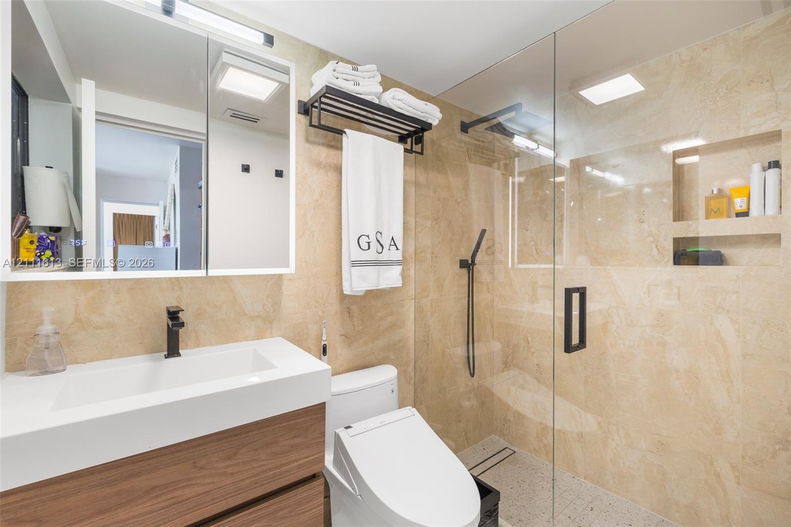 Bathroom featuring a wood-finish floating vanity with an integrated white sink, stone-tiled wall surfaces, a glass-enclosed shower with an integrated 
