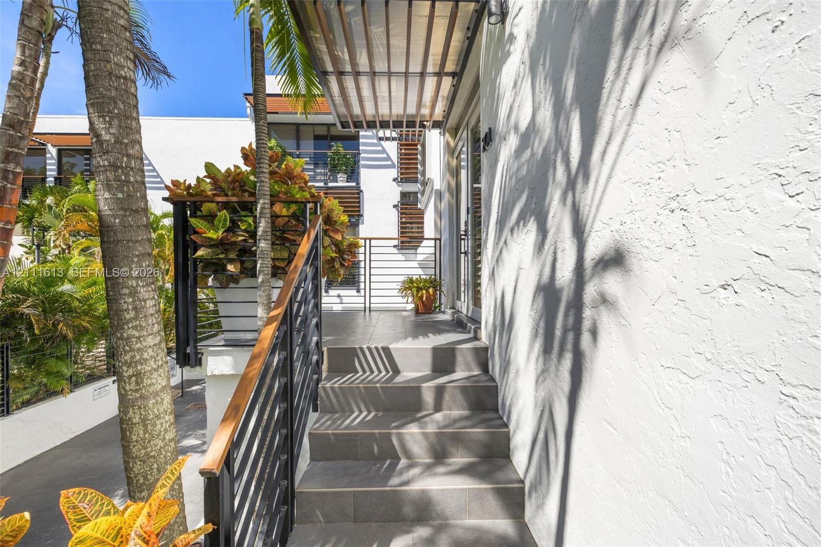 Exterior entryway featuring a modern staircase with tile treads, a wood-accented metal railing, a louvered metal awning, and white textured stucco wal
