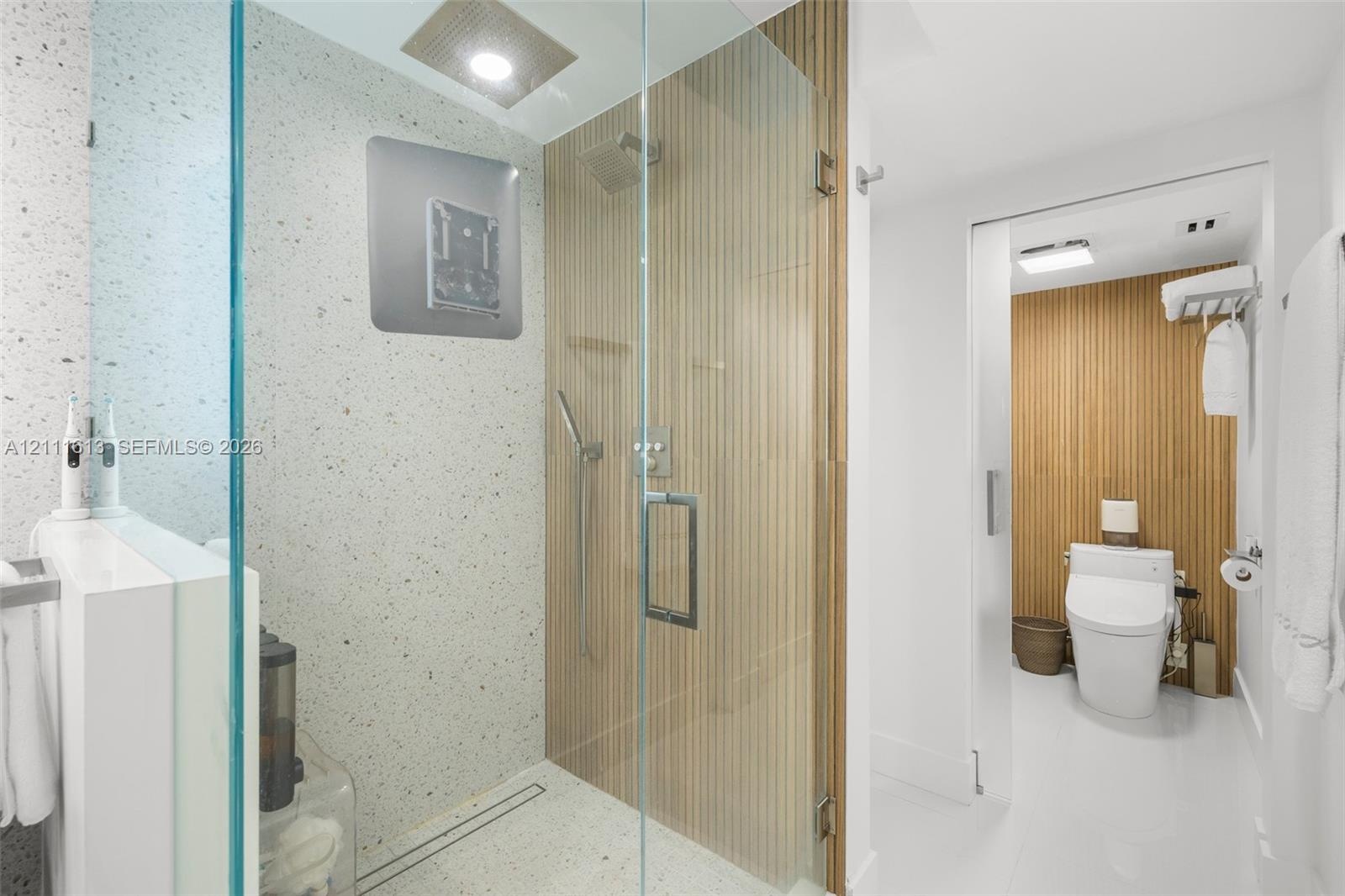 Walk-in shower with terrazzo-style wall and floor tiles, wood-slat accent wall, frameless glass enclosure, rain showerhead, and sliding door entry