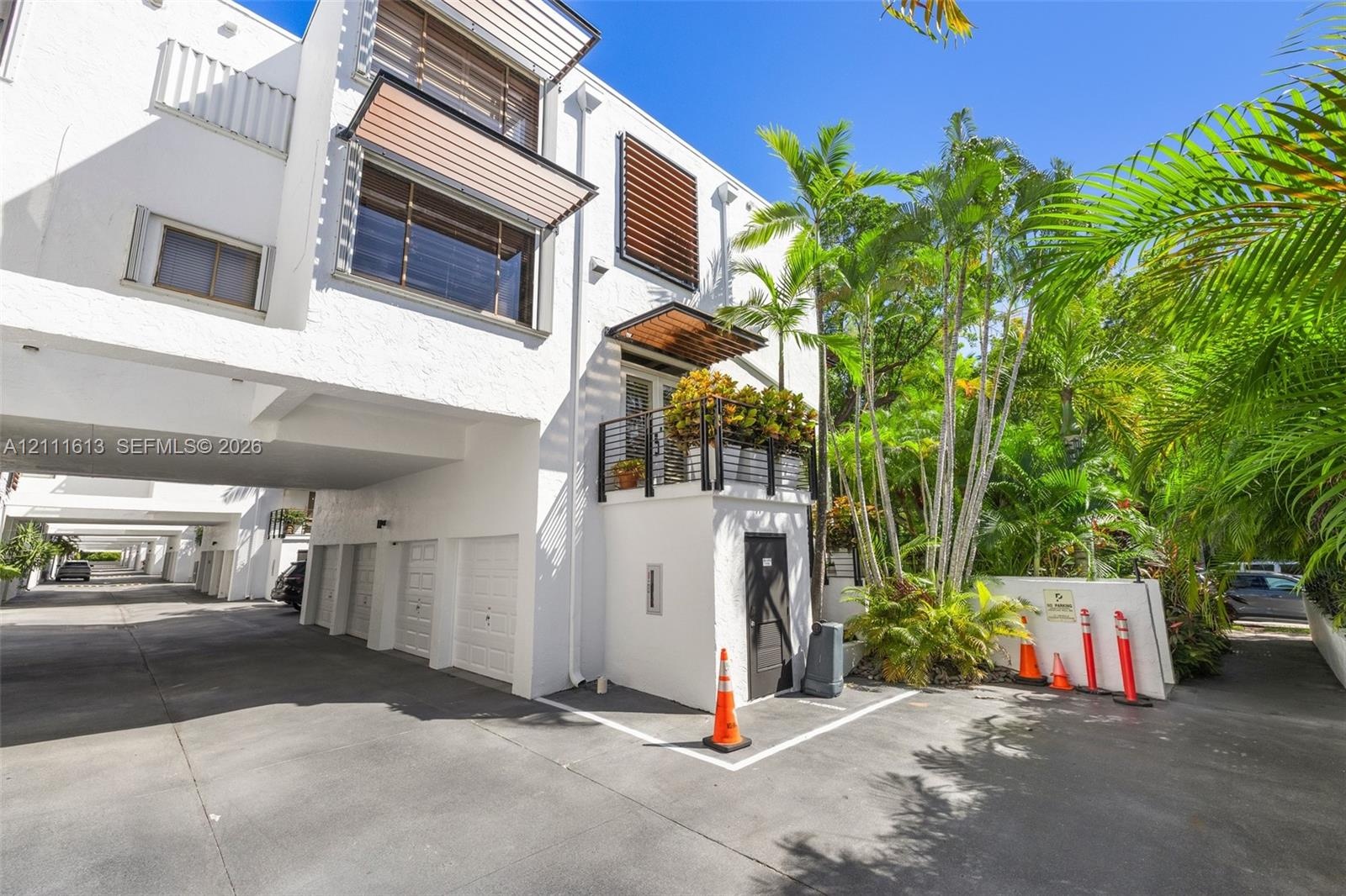 Contemporary multi-level exterior featuring white textured stucco, wood-slat window awnings, elevated balcony with black metal railing, and covered gr