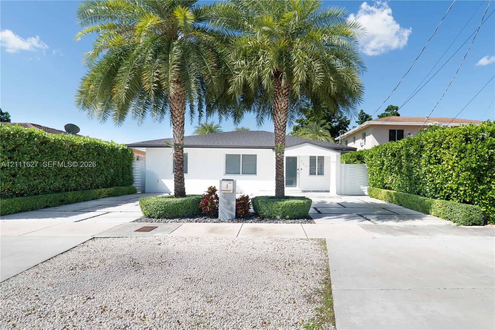 Single-story exterior featuring white stucco siding, a hip roof, expansive concrete paved driveway with integrated stone borders, mature palm trees, a