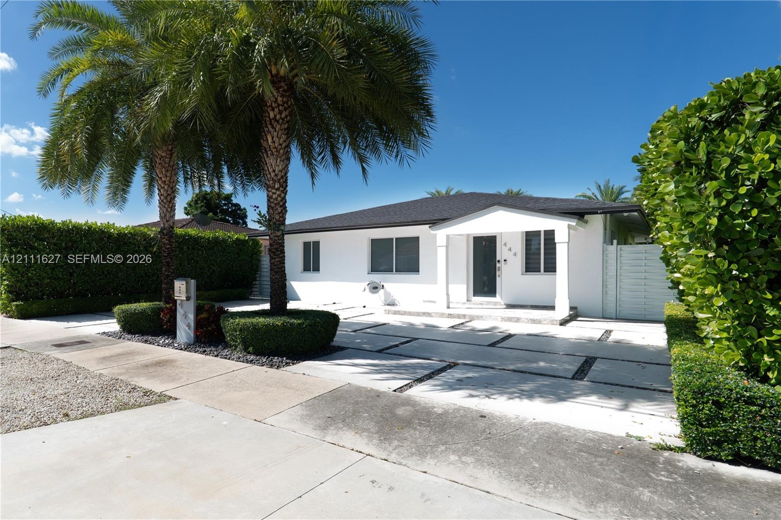 Single-story exterior featuring white stucco siding, a covered entryway with square columns, geometric concrete paver driveway, and mature palm trees