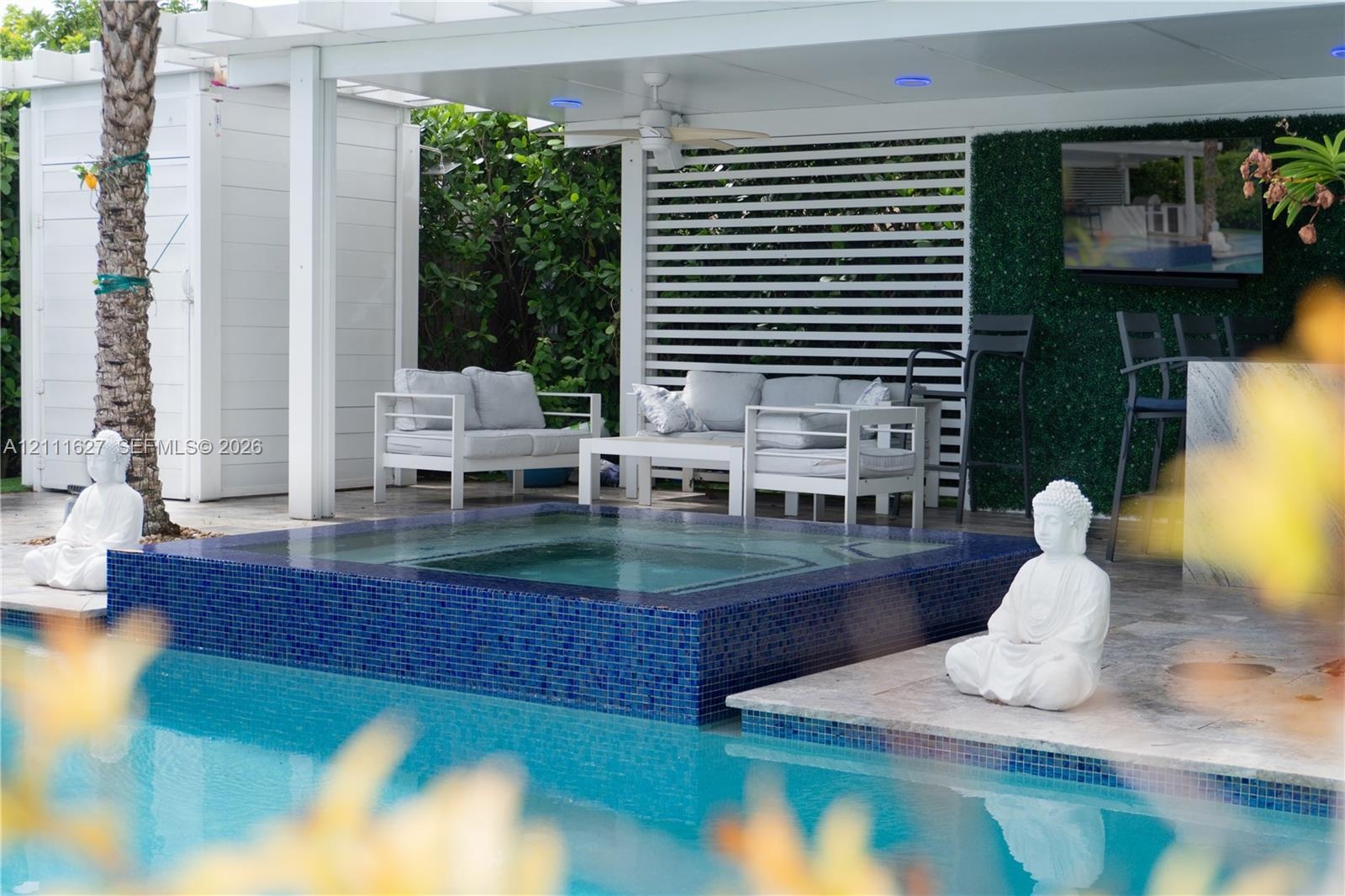 Covered outdoor lounge area featuring a blue mosaic tile spa, stone-finish decking, white slatted privacy screen, and recessed ceiling lighting