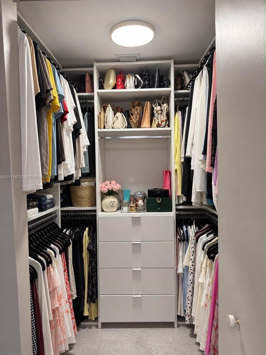 Walk-in closet featuring white built-in shelving, four integrated storage drawers with silver-tone pulls, multiple hanging rods, and a flush-mount ove