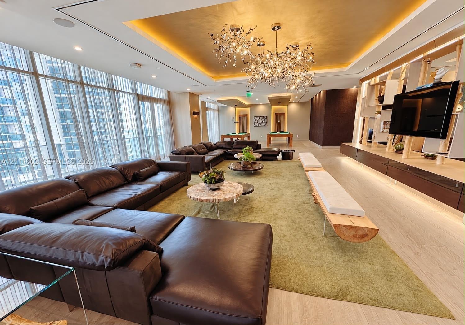 Expansive room featuring floor-to-ceiling windows, gold-leaf tray ceiling, branching metallic statement chandelier, light-toned wood-finish flooring, 