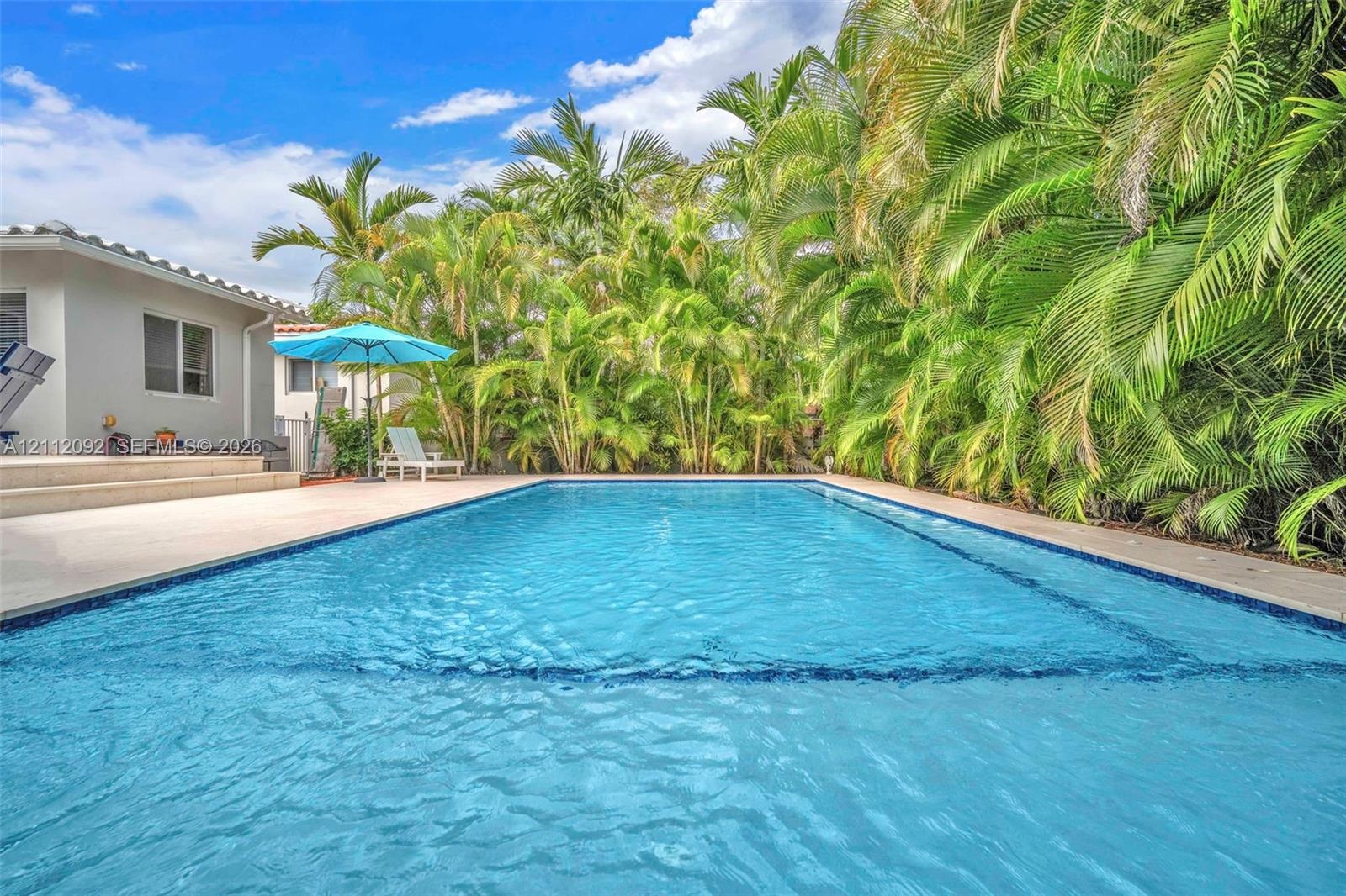 In-ground swimming pool with tile surround, concrete patio, and dense perimeter landscaping