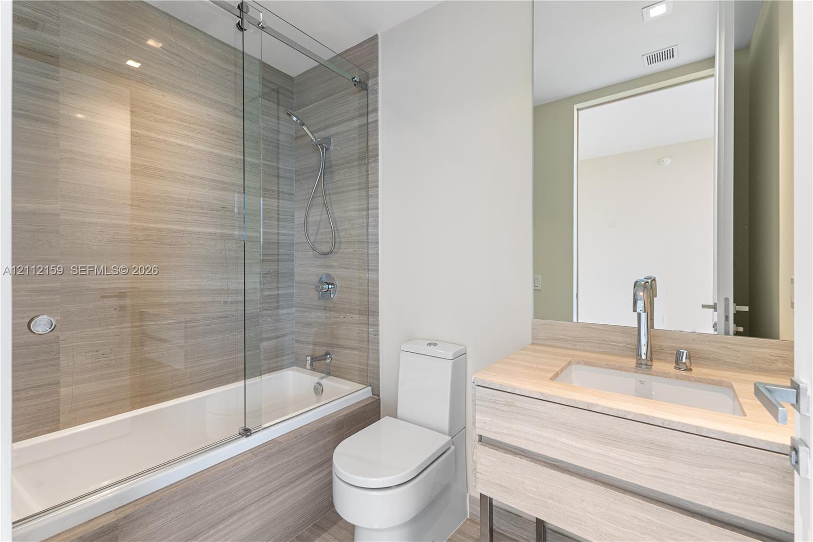 Bathroom featuring a wall-mounted vanity with integrated sink, glass-enclosed tub with handheld shower fixture, and horizontal tile wall surrounds