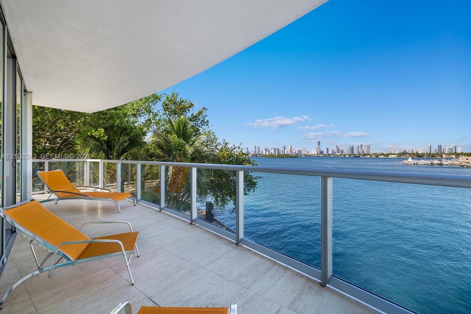 Covered balcony featuring stone flooring and a glass railing with metal framing, overlooking the waterfront and city skyline
