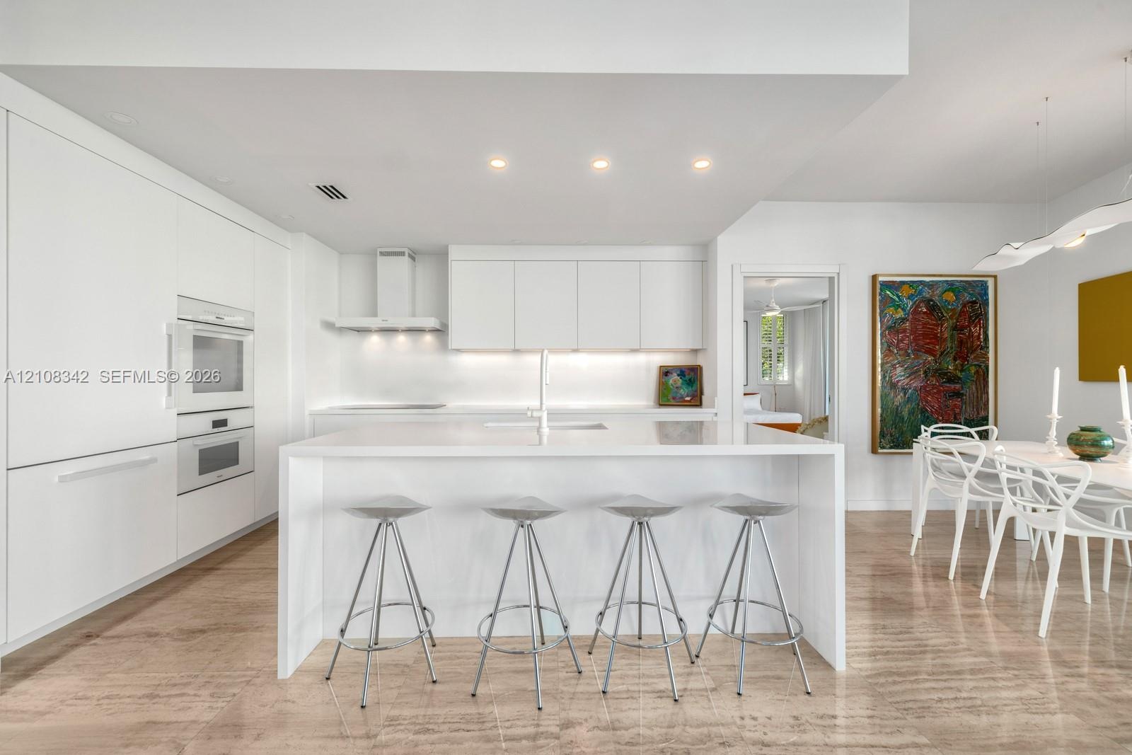 Modern kitchen featuring white flat-panel cabinetry, a central island with a solid-surface countertop, built-in double ovens, and polished stone floor