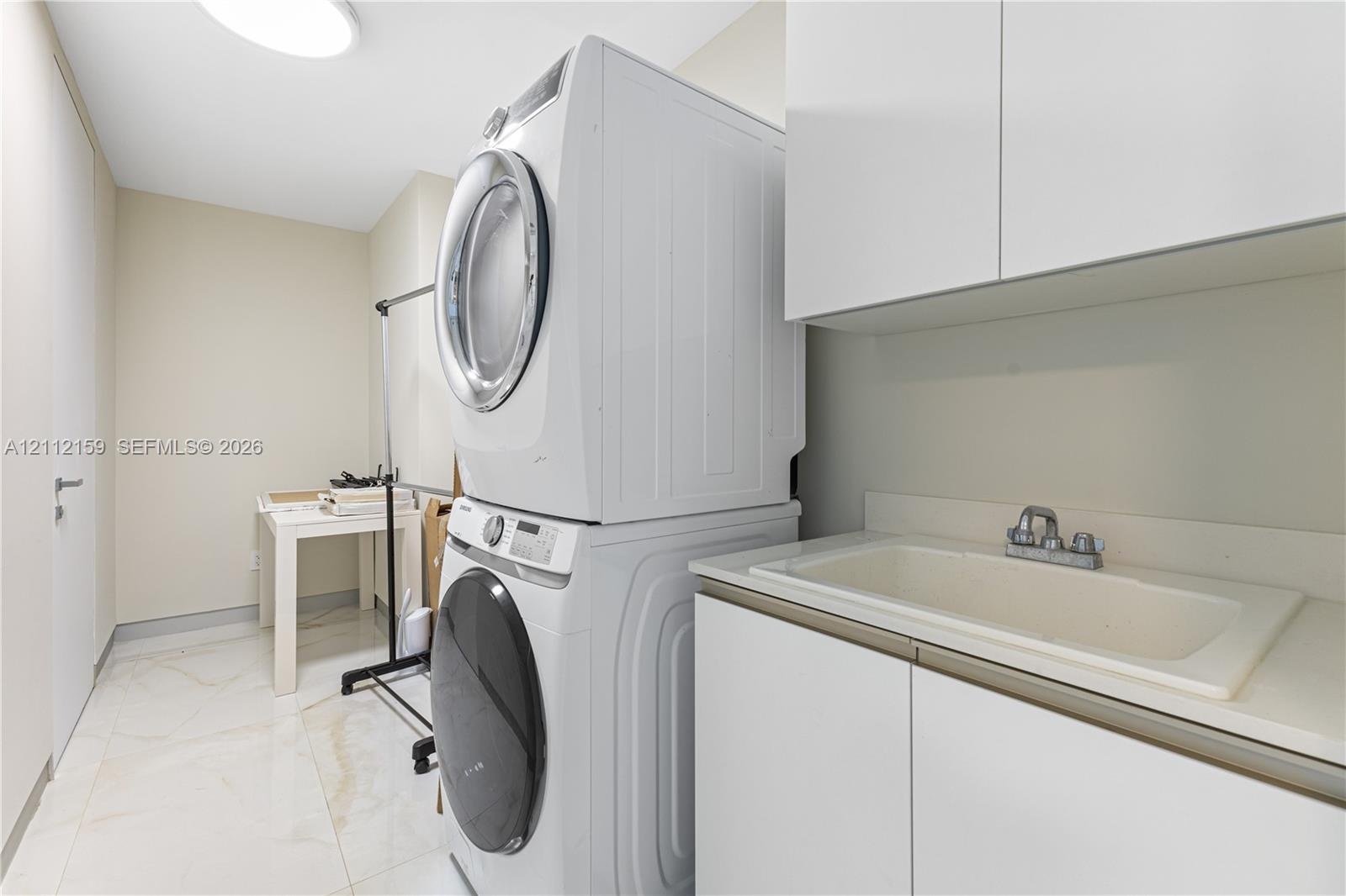 Laundry space featuring stacked front-load laundry appliances, white cabinetry with integrated sink, and polished tile flooring