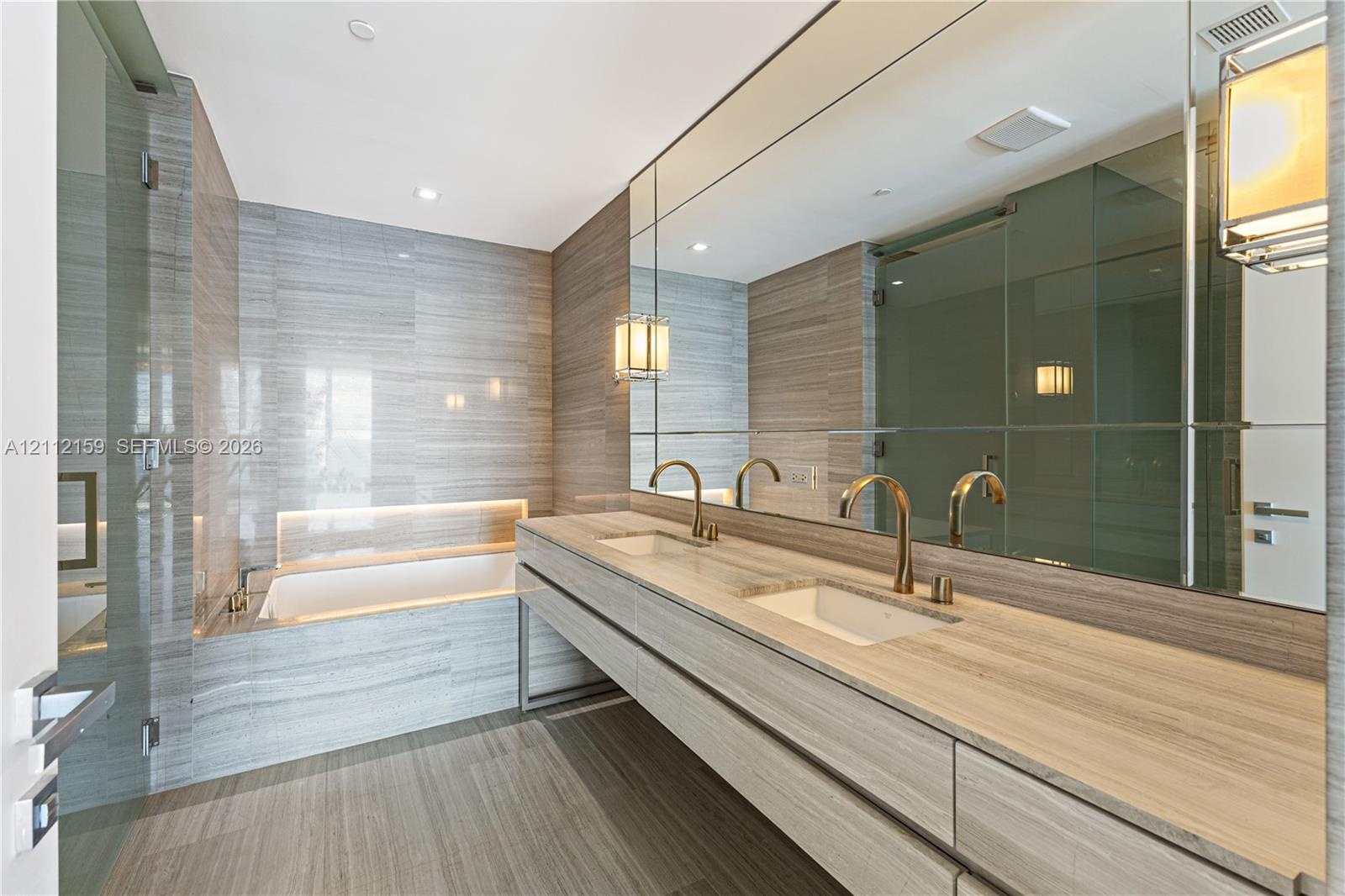 Dual-vanity cabinet featuring stone-topped surfaces, undermount sinks, brass-toned fixtures, floor-to-ceiling tiled walls, and a recessed soaking tub 