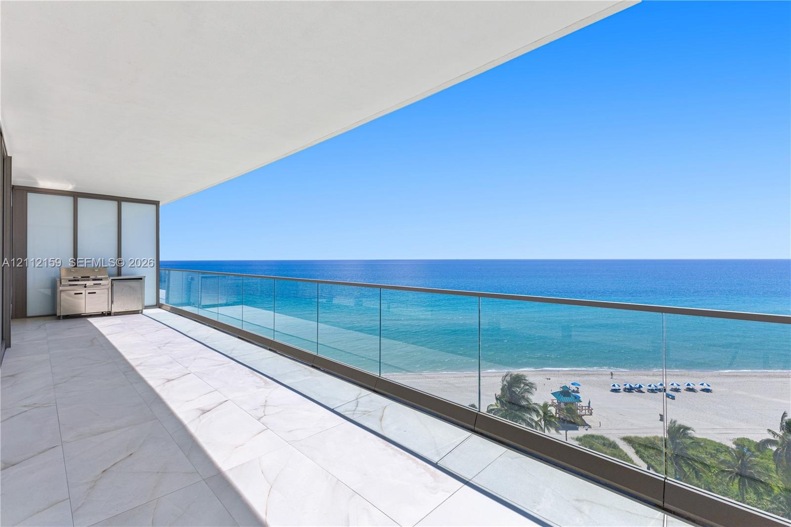 Spacious covered balcony featuring large-format stone tile flooring, a glass railing, and a built-in stainless steel outdoor kitchen station