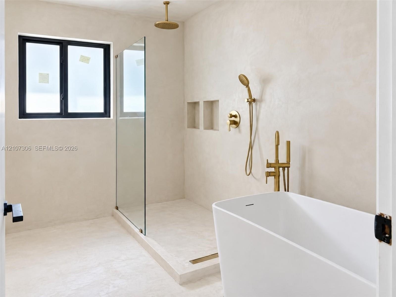 Freestanding soaking tub with brass fixtures, glass-enclosed walk-in shower, dual recessed wall niches, and matte-finish plaster wall treatment