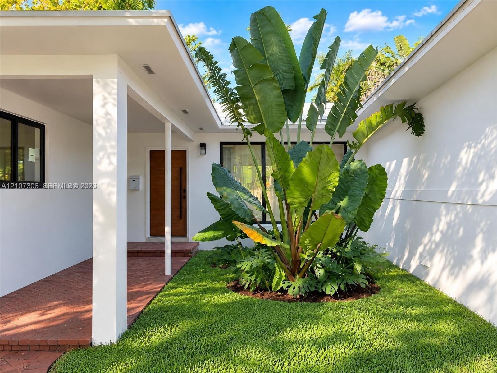 Courtyard entryway featuring a red brick floor, white stucco exterior walls, a natural wood-finish front door, and mature tropical landscaping