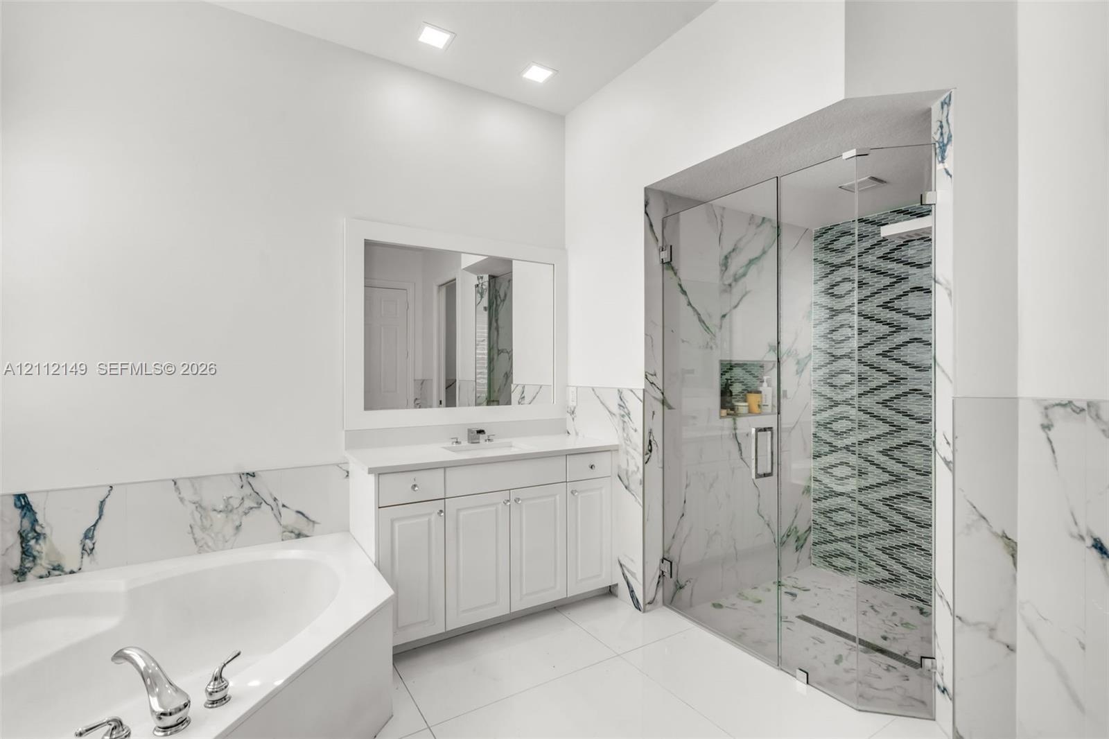 White vanity with integrated basin, framed wall mirror, soaking tub with deck-mounted faucet, walk-in shower with glass enclosure, and patterned tile 