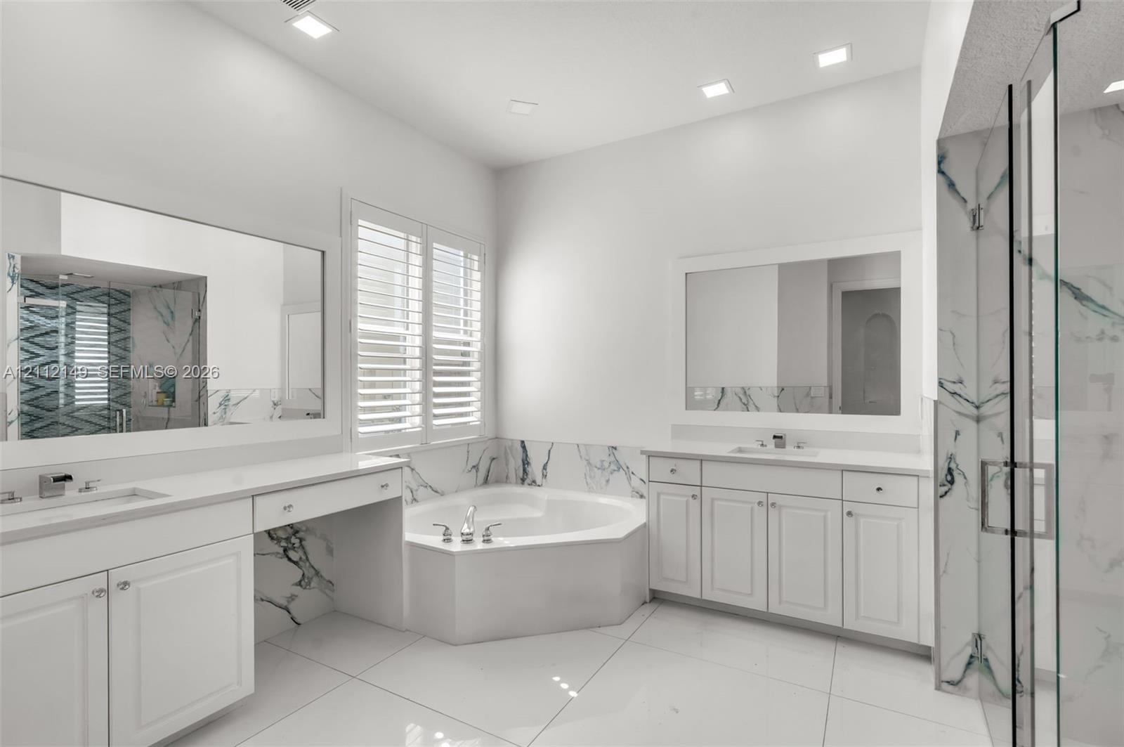 Bathroom featuring white cabinetry, integrated vanity, dual-basin sinks, jetted soaking tub, and stone-accented shower enclosure with glass door