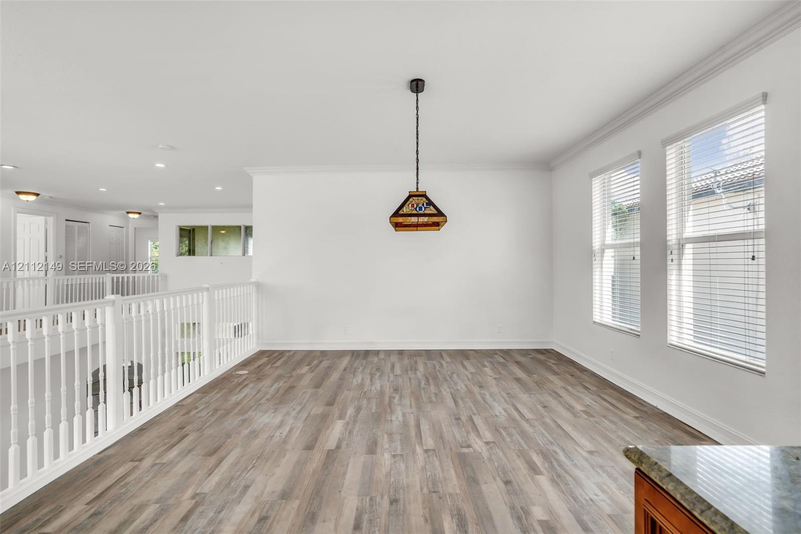 Open space featuring wood-finish flooring, white walls, crown molding, double windows with horizontal blinds, and a suspended geometric pendant light 
