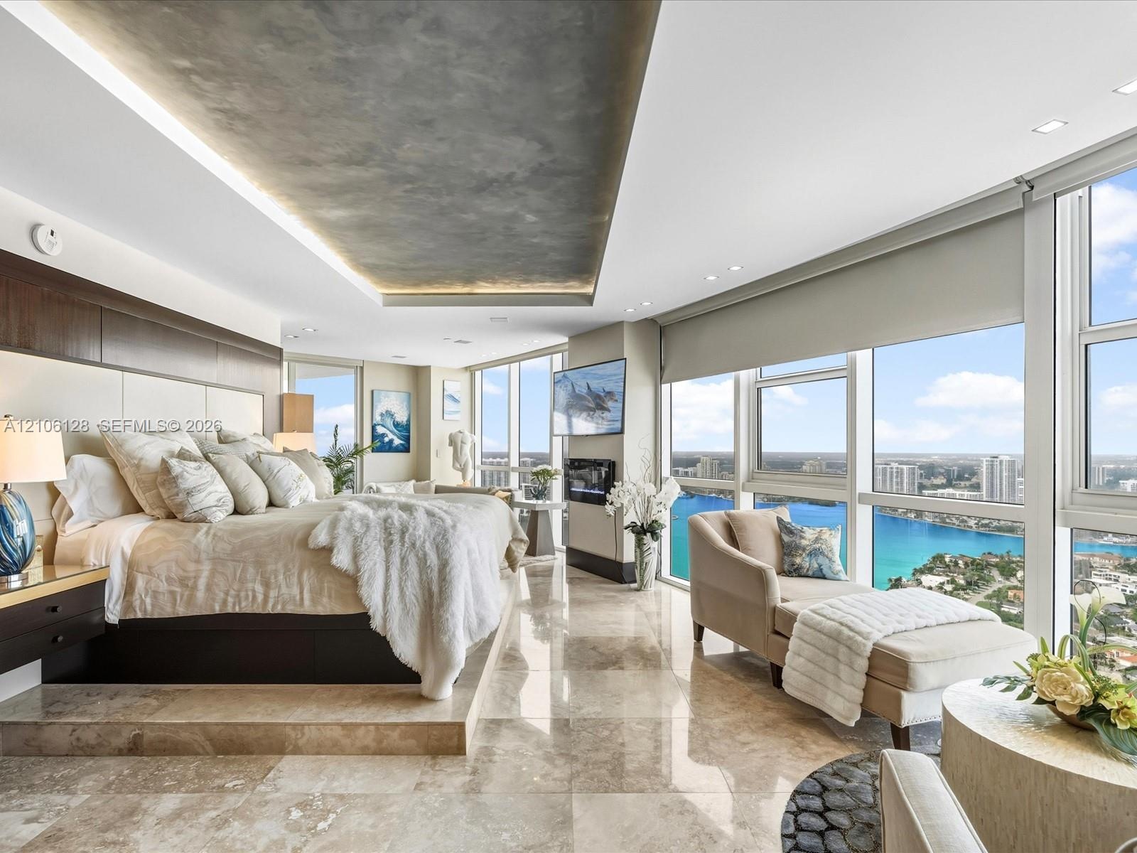 Floor-to-ceiling windows, stone tile flooring, tray ceiling with recessed lighting, wall-mounted fireplace, and elevated platform bed base