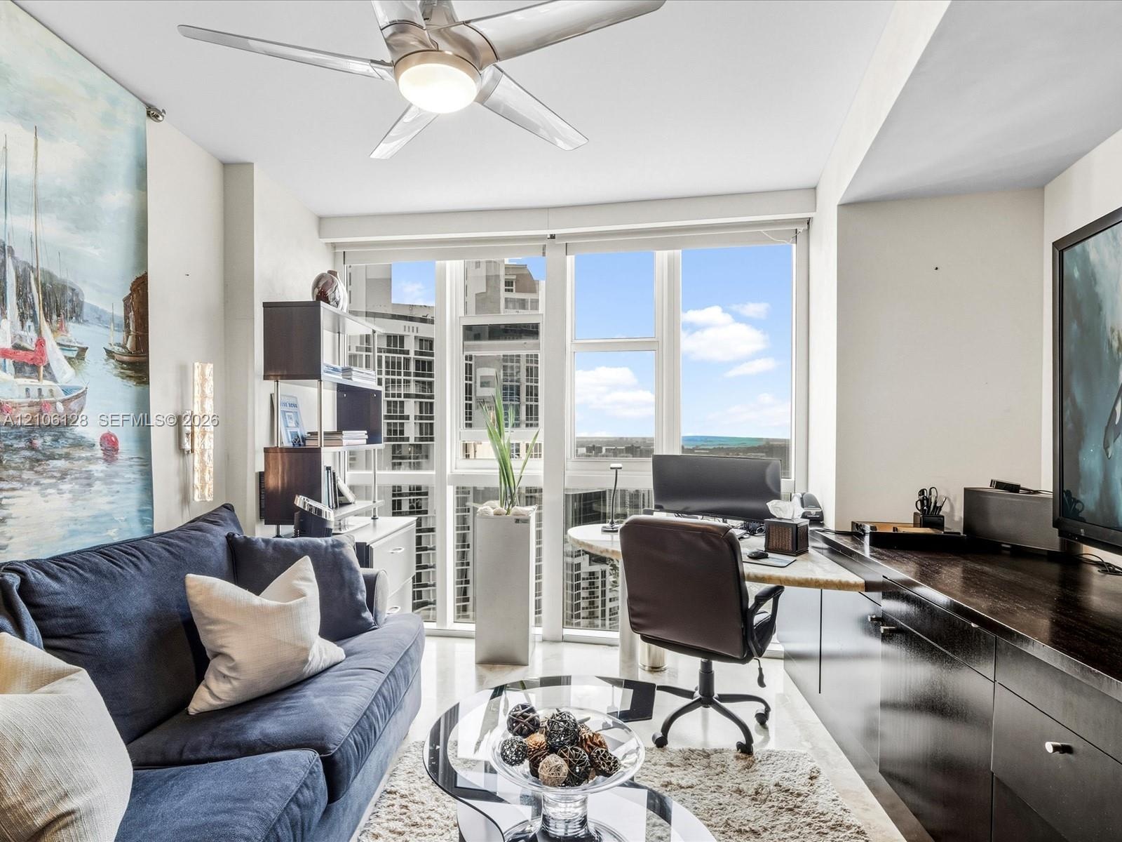 Floor-to-ceiling windows, built-in dark cabinetry with integrated desk space, and a ceiling-mounted light fixture with fan