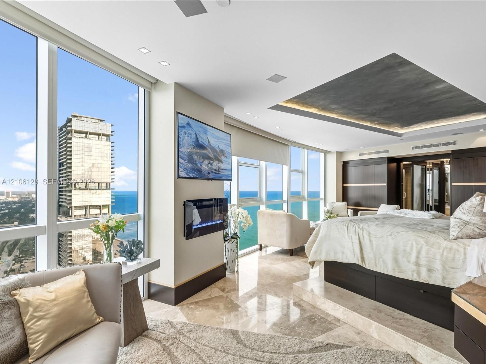 Floor-to-ceiling windows featuring ocean scenery, recessed ceiling detailing with integrated lighting, built-in wall-mounted electric fireplace, raise