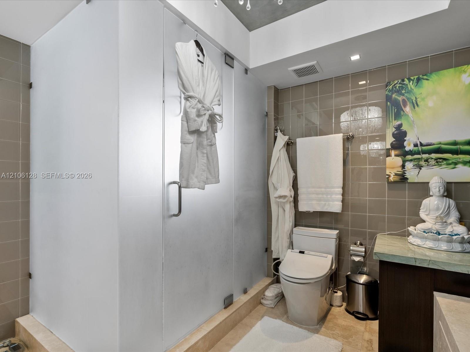 Walk-in shower enclosure featuring frosted glass panels, neutral-toned wall tile, modern dual-flush toilet, and stone-surface countertop