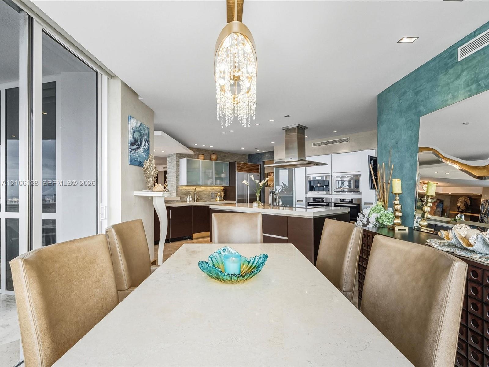 Open-concept interior featuring a stone-topped dining area, a tiered crystal chandelier, custom flat-panel cabinetry, a stainless steel chimney hood, 