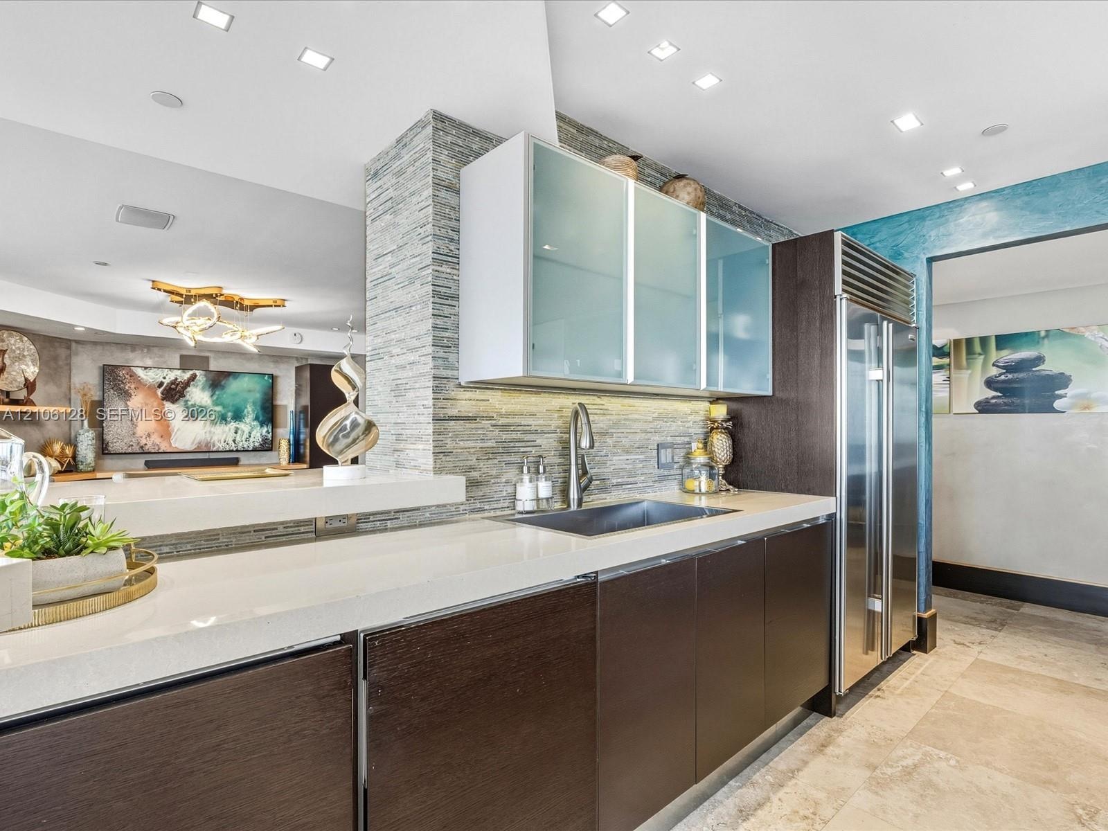 Modern kitchen featuring dark wood-finish cabinetry, frosted glass upper cabinets, a light-toned countertop, a built-in stainless steel refrigerator, 