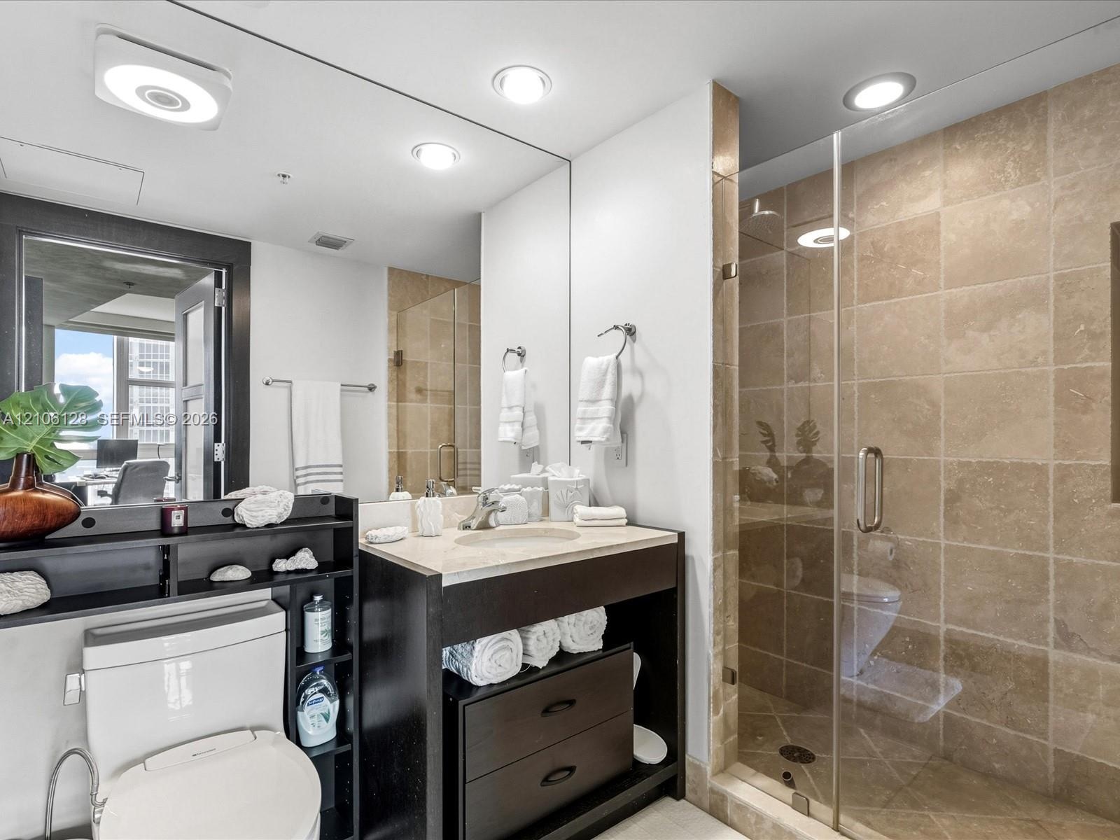 Bathroom featuring a vanity with stone countertop, under-mount sink, dark wood-finish cabinetry, full-width mirror, and a walk-in shower with tile sur