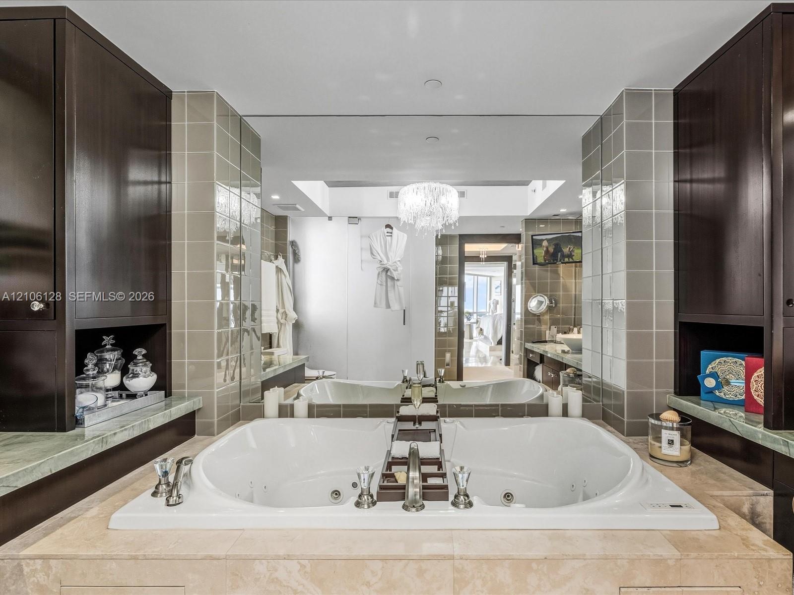 Jetted soaking tub with deck-mounted chrome fixtures, flanked by dark wood cabinetry, integrated stone countertops, and full-wall mirror accented by t