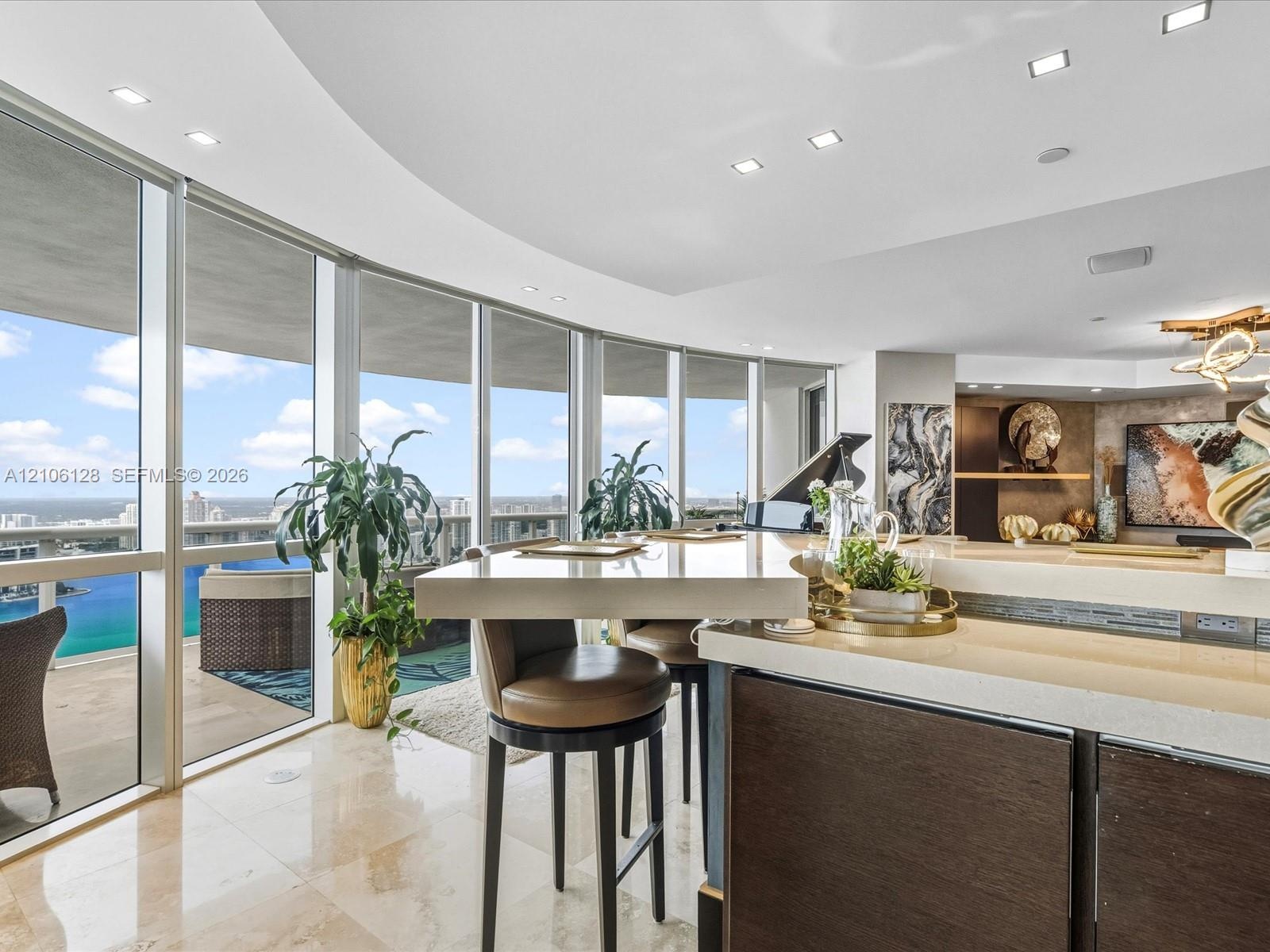 Curved floor-to-ceiling windows, light-toned stone flooring, recessed ceiling lighting, and a multi-level kitchen island with a stone countertop