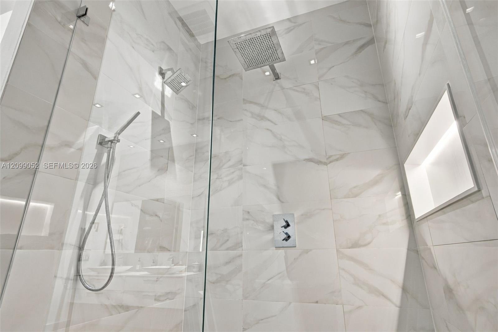 Walk-in shower featuring large-format light veined tile, frameless glass enclosure, ceiling-mounted rainfall showerhead, adjustable handheld shower wa