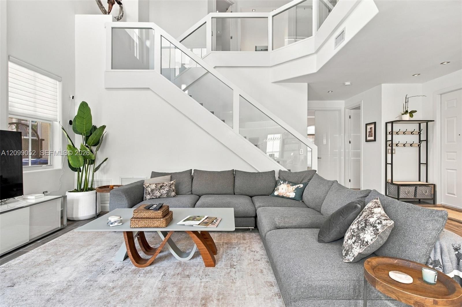 Double-height living area featuring a custom glass and white-frame staircase railing, recessed lighting, and large windows with horizontal blinds